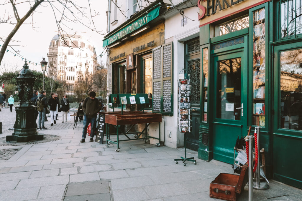 Außenansicht von Shakespeare and Company Paris mit Blick von französischen Gebäuden im Hintergrund und Menschen, die sich Bücher ansehen.